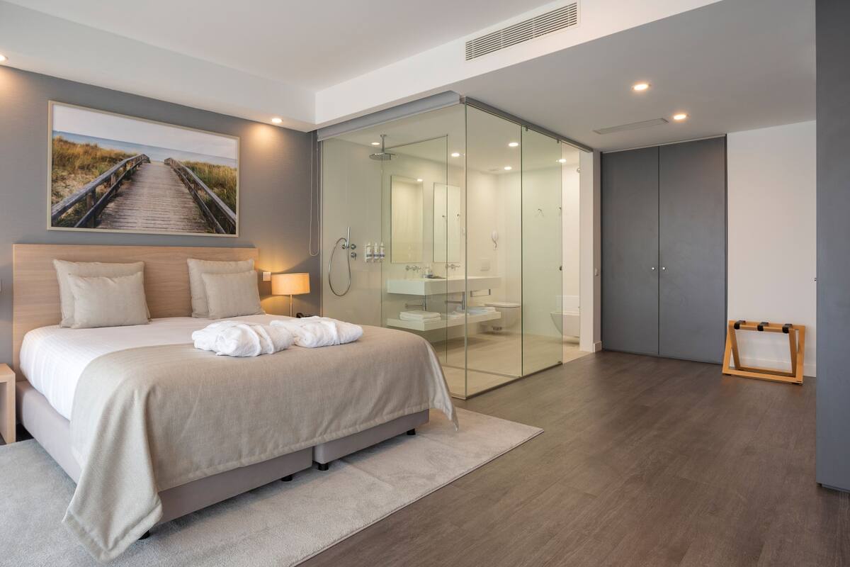 A modern bedroom with a large bed, bedside lamps, a glass shower enclosure, and a view of a beach pathway.