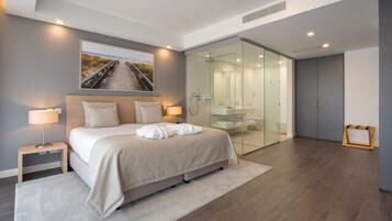 A modern bedroom with a large bed, bedside lamps, a glass shower enclosure, and a view of a beach pathway.