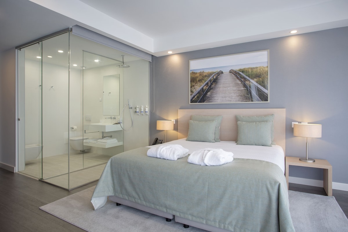 A modern hotel room with a glass-enclosed shower, a large bed with two pillows, and a painting of a beach walkway on the wall.
