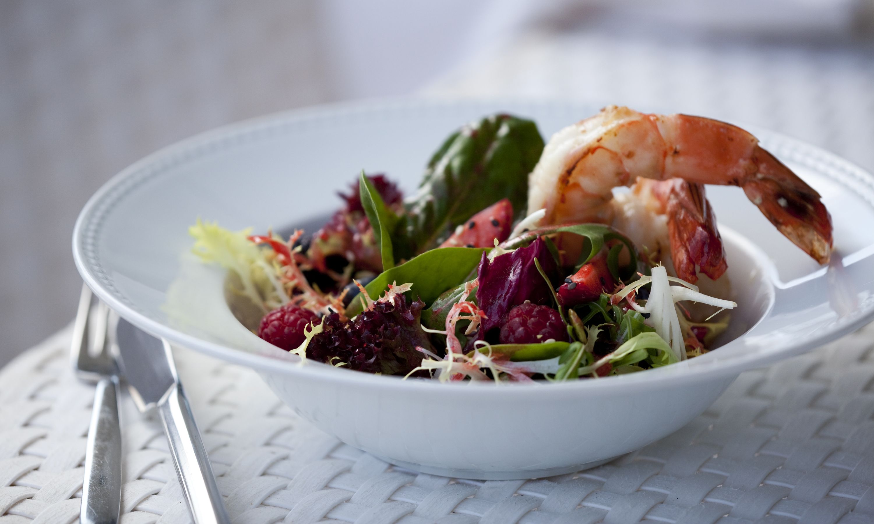 Un saladier blanc contenant une salade composée de différentes feuilles vertes, de baies rouges et d’une crevette, le tout posé sur une nappe à motifs tissés.