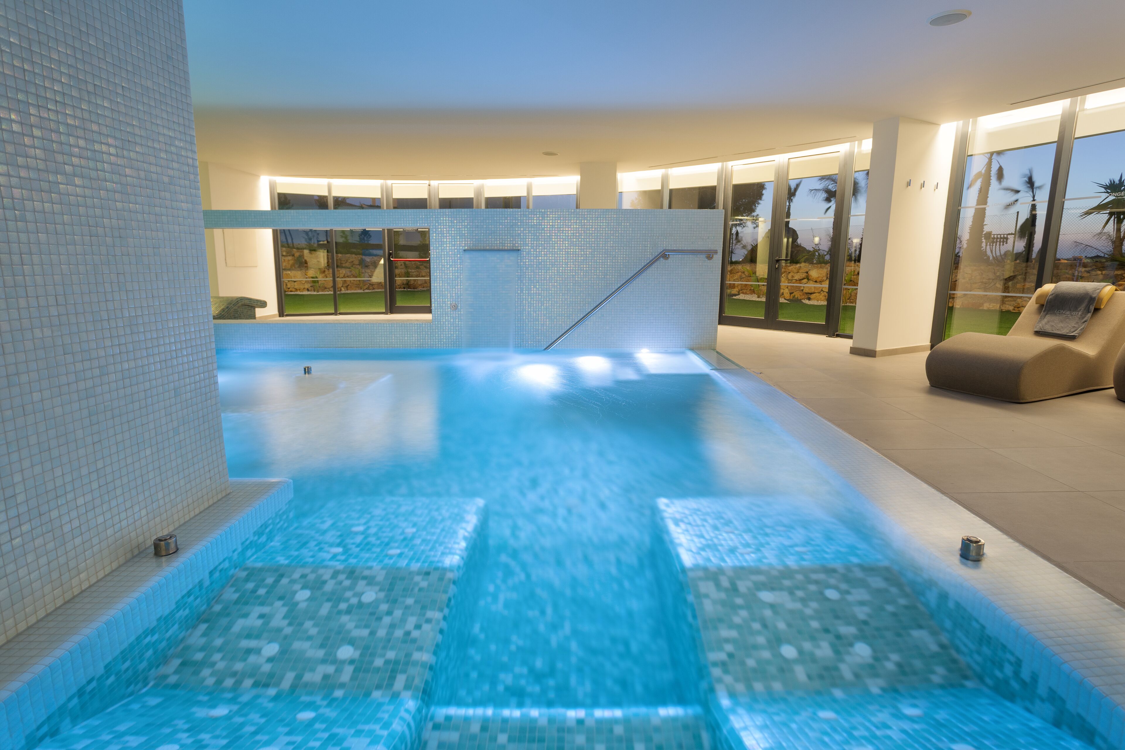 Una piscina interior moderna con paredes de vidrio transparente, suelo de baldosas y un sillón de sol.