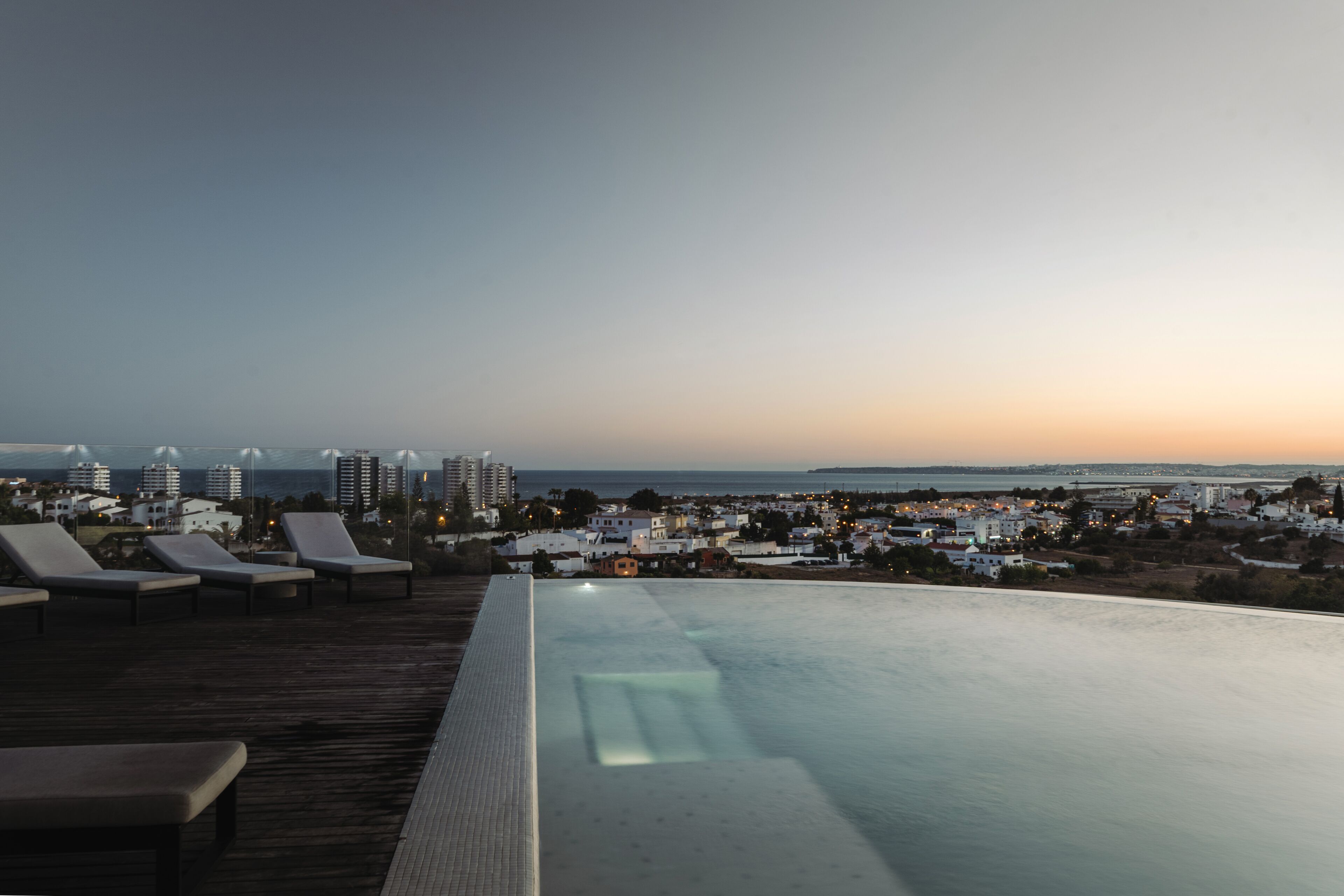 A rooftop infinity pool with lounge chairs and a city view at dusk.