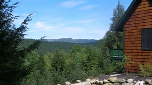 A wooden cabin with a stone pathway leading to a green bench, surrounded by dense forest and mountains in the background.