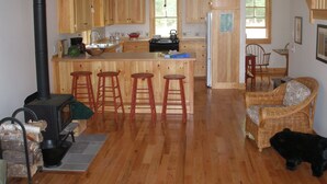A spacious kitchen with wooden cabinets, a central island with stools, a black stove, and a dining area with a wicker chair and a black dog.
