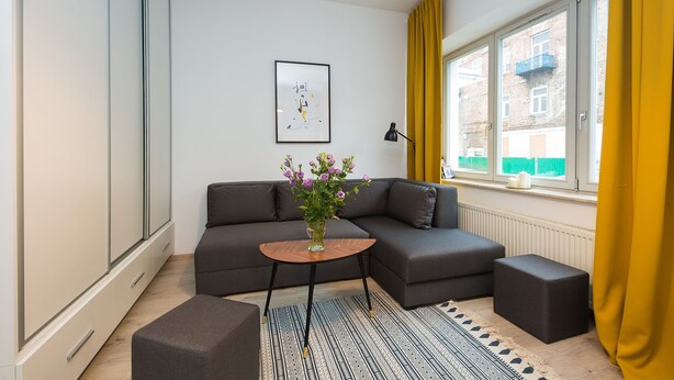 A modern living room with a grey sofa, a small round table with a vase of flowers, a black floor lamp, and a window with yellow curtains. - ShortStayPoland Środkowa (B48) (Warsaw)
