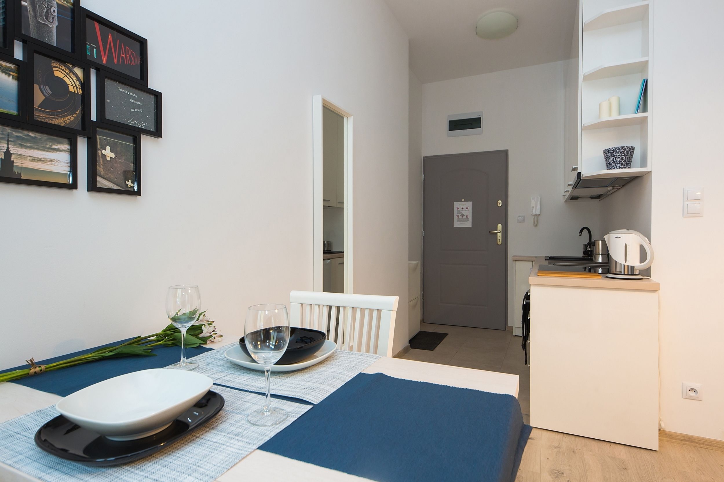 A compact room with a dining table set for two, a kitchenette, and a door leading to another room.