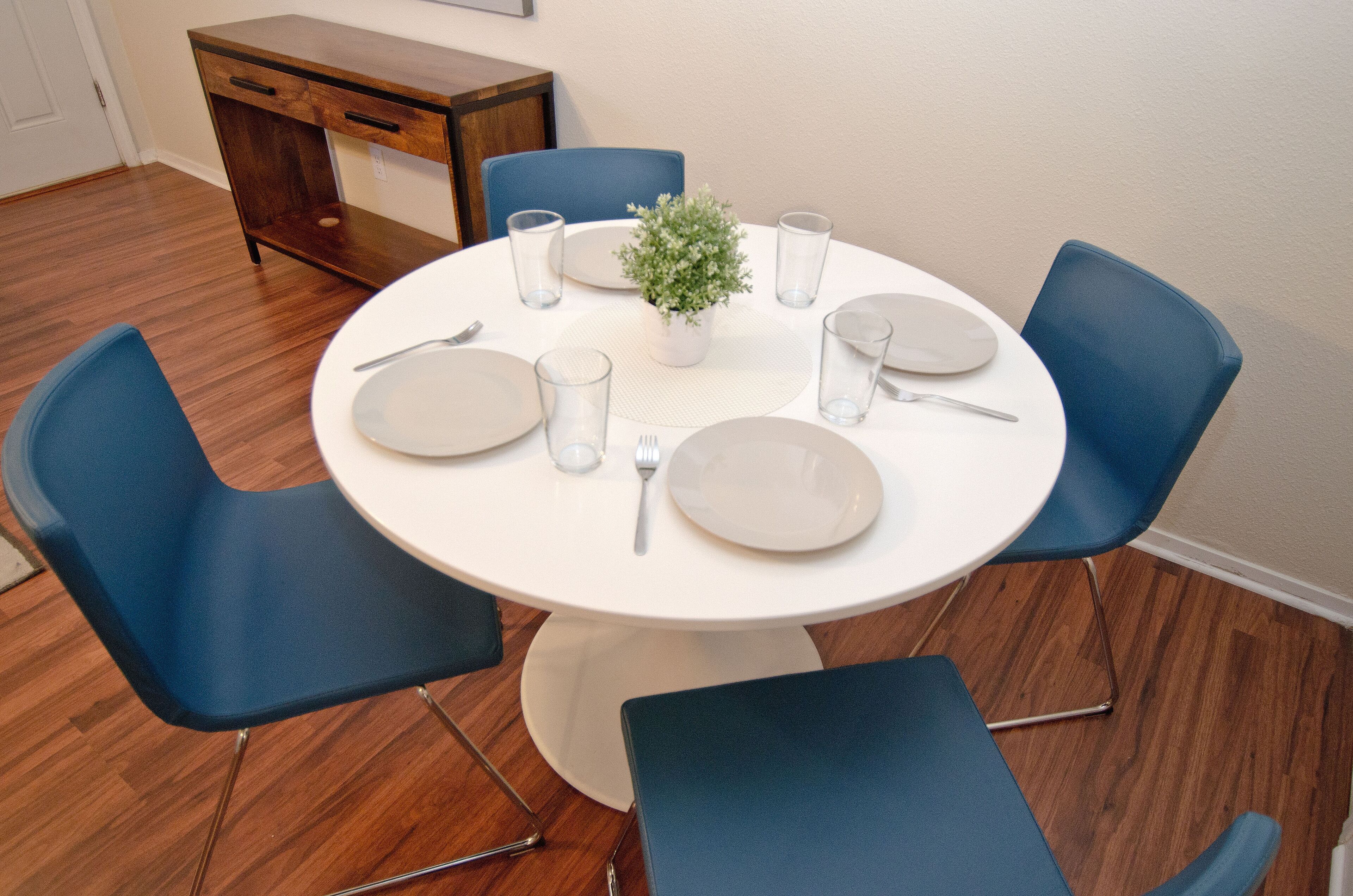 A round dining table with six blue chairs, white plates, cutlery, and glasses, centered on a wooden floor with a small potted plant.
