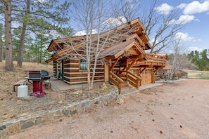 A rustic log cabin with a porch, a grill, and a stone retaining wall.