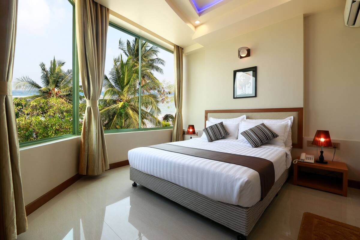 Ein Hotelzimmer mit einem großen Bett, einem Nachttisch, einer Lampe und Blick auf Palmen und das Meer.