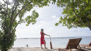 Een strandscène met een persoon in een rode jurk en hoed, een houten stoel en een gitaar.