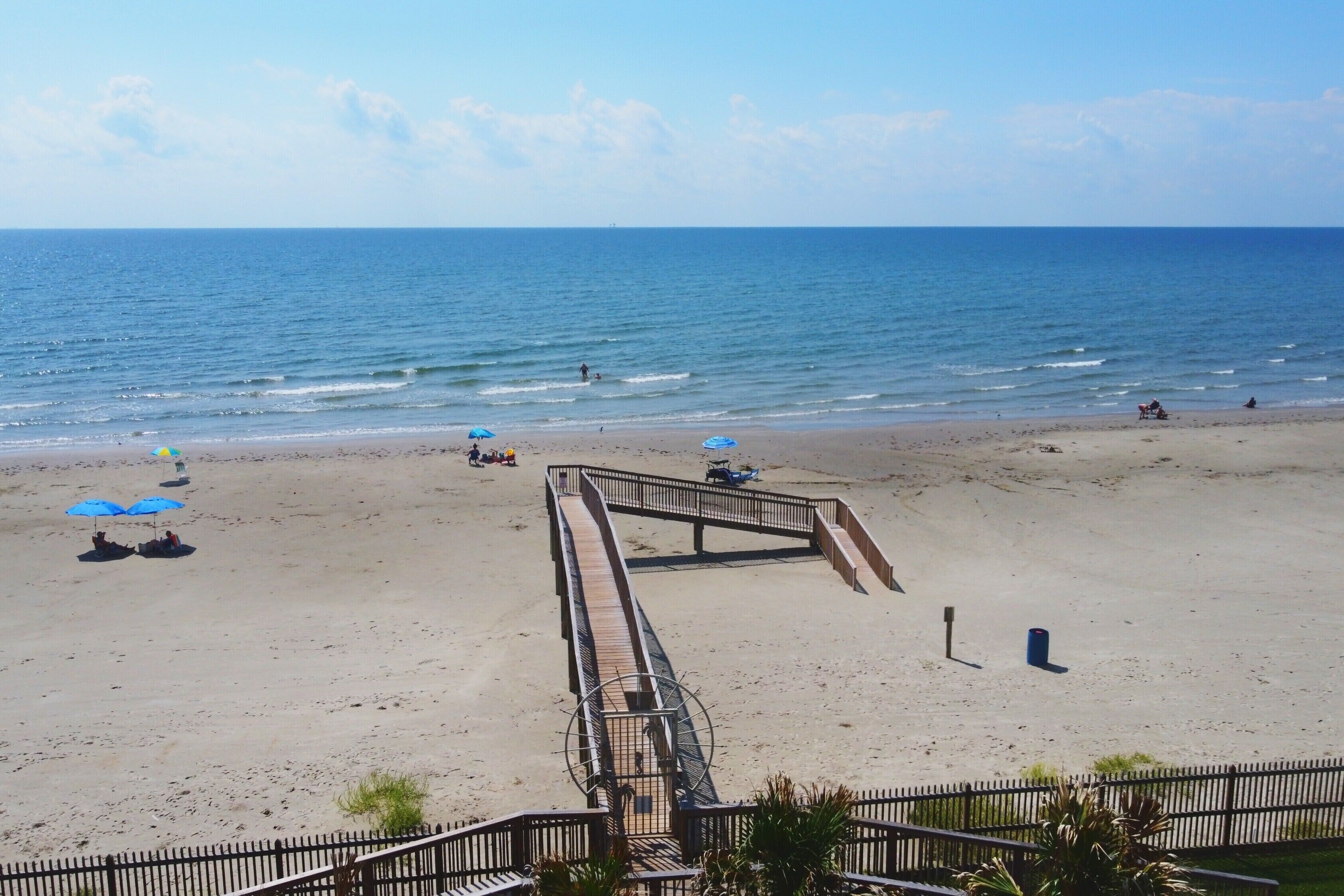 A beach with a wooden boardwalk leading to the water, umbrellas, and a few people.