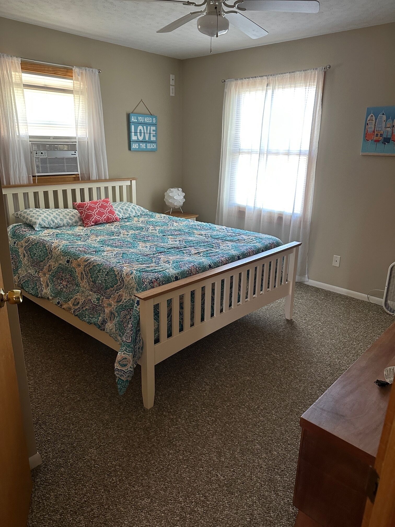 A bedroom with a bed, a ceiling fan, a window with curtains, a wall clock, and a small table with a lamp.