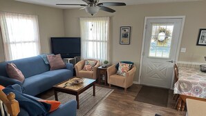 A living room with a blue sofa, a small table, two armchairs, a television, and a dining area with a patterned tablecloth.