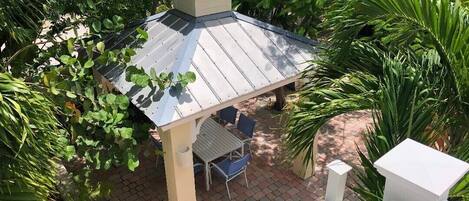 A covered patio area with a table and chairs, surrounded by lush greenery.