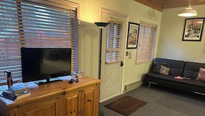 A living room with a TV on a wooden cabinet, a sofa, and a door with blinds. - Cozy Cabin Minutes from Glacier's Two Medicine Valley (East Glacier)