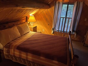 Un dormitorio de cabaña acogedor con una cama, una mesita de noche, una lámpara y un ventanal con cortinas.