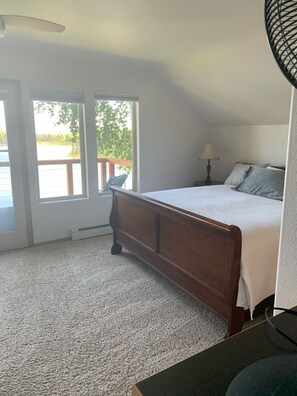 A bedroom with a wooden bed, a nightstand, a lamp, and a view of a lake through large windows. - Vacation Home On The Secluded Shores Of Loon Lake (Soldotna)