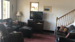 Ein Wohnzimmer mit Holzboden, einem Flachbildfernseher auf einem Holzbodenständer, Ledersofas, einem roten Teppich und einer Treppe.