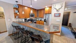 A kitchen with wooden cabinets, a stainless steel refrigerator, a bar area with stools, and a mounted deer head decoration. - Luxury Condo w/ FREE WiFi, Parking, Heated Pool, Hot Tubs, Skier Shuttles (Avon)