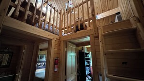 Un interior de madera con un entrepiso, una puerta que da a otra habitación y una estantería con objetos varios.