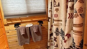 Interior de una cabaña de madera con una ventana cubierta por persianas, un banco de madera y una cortina con motivos naturales.