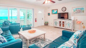 A living room with a TV, a ceiling fan, and a view of the ocean.