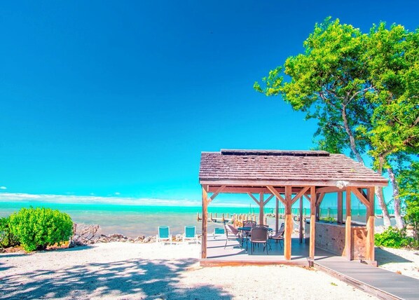 A wooden gazebo with a tiled roof, surrounded by beach chairs and tables, overlooking a clear blue ocean.