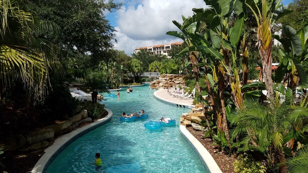 Zona de piscina amb cascada, envoltada de vegetació abundant i un edifici al fons.