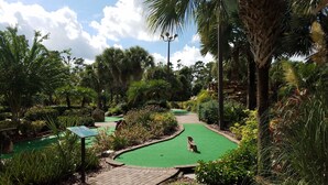 Camp de golf amb una zona de green, envoltat de vegetació abundant i palmeres.