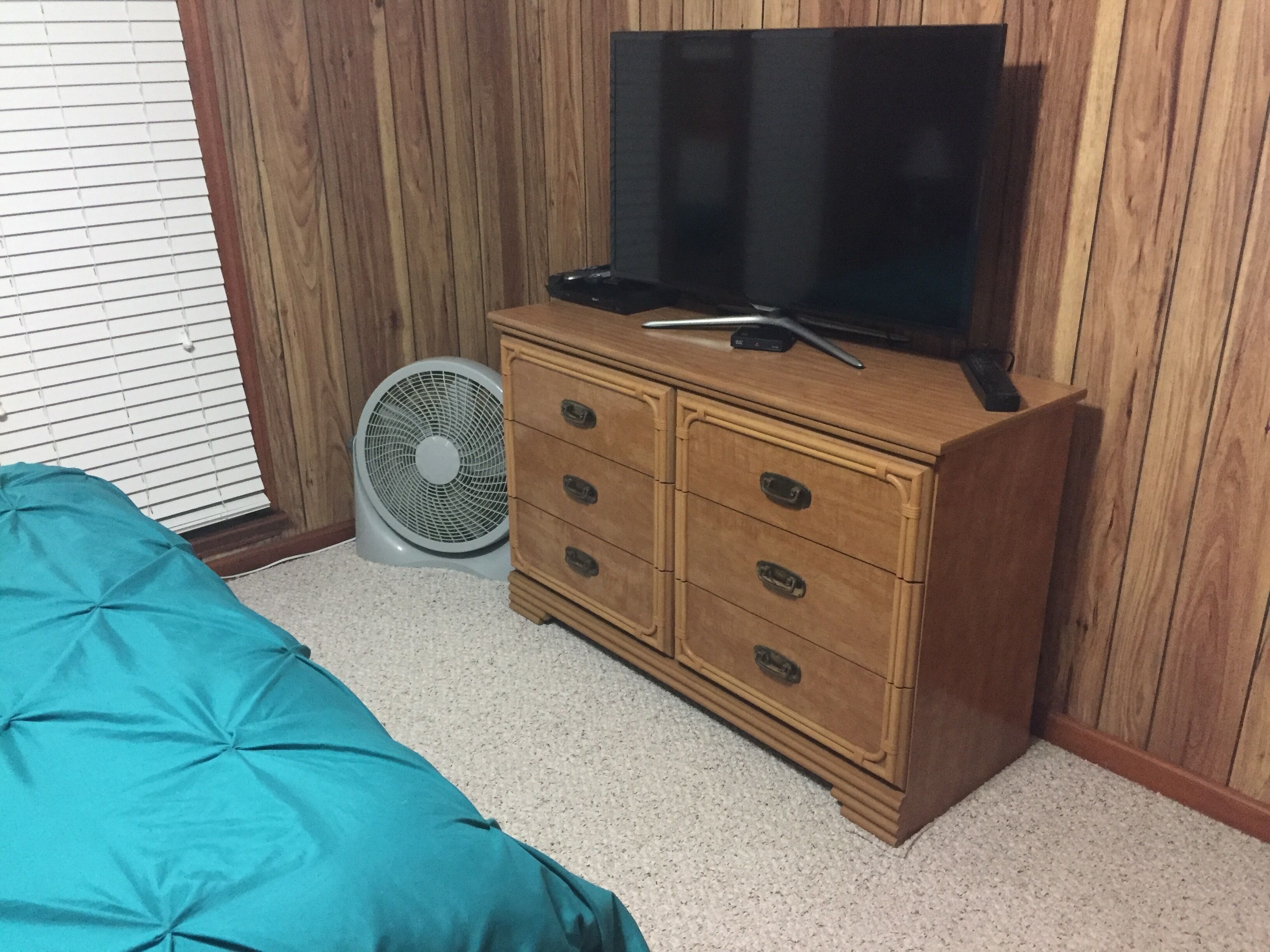 Una camera da letto con un comò in legno, un televisore sopra, un ventilatore e una coperta letto color petrolio.