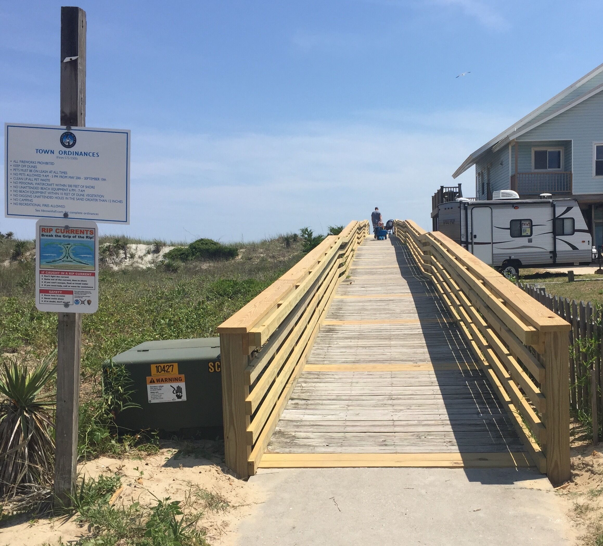 Une promenade en bois mène à une zone de plage où un panneau indique les règlements municipaux et met en garde contre les courants de contre-rivage.