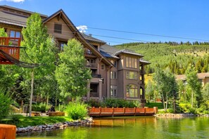 Un chalet à plusieurs étages doté d’un grand balcon, entouré d’une végétation luxuriante et bordé d’un lac.