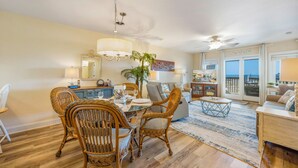 A spacious living area with a dining table, wicker chairs, a large mirror, and a view of the beach through the sliding doors. - Semi-Oceanfront Condo w/ Resort Pool, Elevator, Private Sundeck. Top Notch Stay! (Rodanthe)