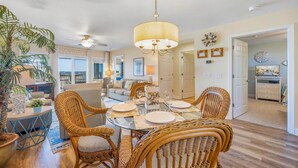 A dining area with wicker chairs, a glass table, and a chandelier. - Semi-Oceanfront Condo w/ Resort Pool, Elevator, Private Sundeck. Top Notch Stay! (Rodanthe)