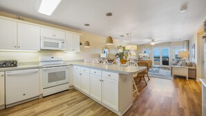 A spacious kitchen with white cabinetry, a central island, and a dining area with wicker chairs. - Semi-Oceanfront Condo w/ Resort Pool, Elevator, Private Sundeck. Top Notch Stay! (Rodanthe)