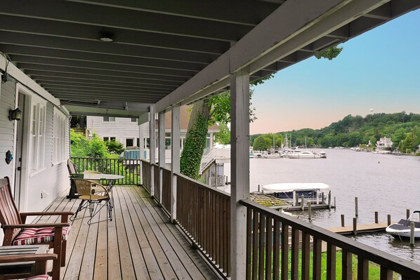 A wooden deck with chairs and a table, overlooking a calm river with boats and houses in the background.