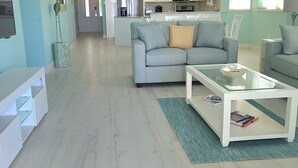 A modern living room with a grey sofa, a glass coffee table, and a kitchen in the background. - YOU'VE Arrived! Beachfront 3BR condo w/ Pool-Relax... North Side near Rum Point (Old Man Bay)
