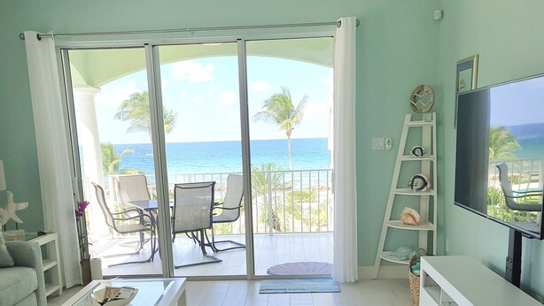 A living room with a view of the ocean, a balcony with chairs, a TV, and a bookshelf. - YOU'VE Arrived! Beachfront 3BR condo w/ Pool-Relax... North Side near Rum Point (Old Man Bay)
