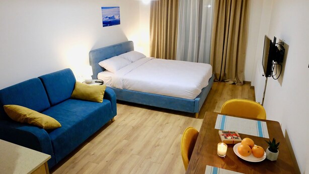 A hotel room with a bed, a blue sofa, a wooden dining table with a plate of oranges, a small potted plant, and a wall-mounted TV. - GUDASKI Apartments (Kazbegi)