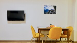 A dining area with a wooden table, four yellow chairs, a flat-screen TV mounted on the wall, and a framed picture of a mountain landscape. - GUDASKI Apartments (Kazbegi)