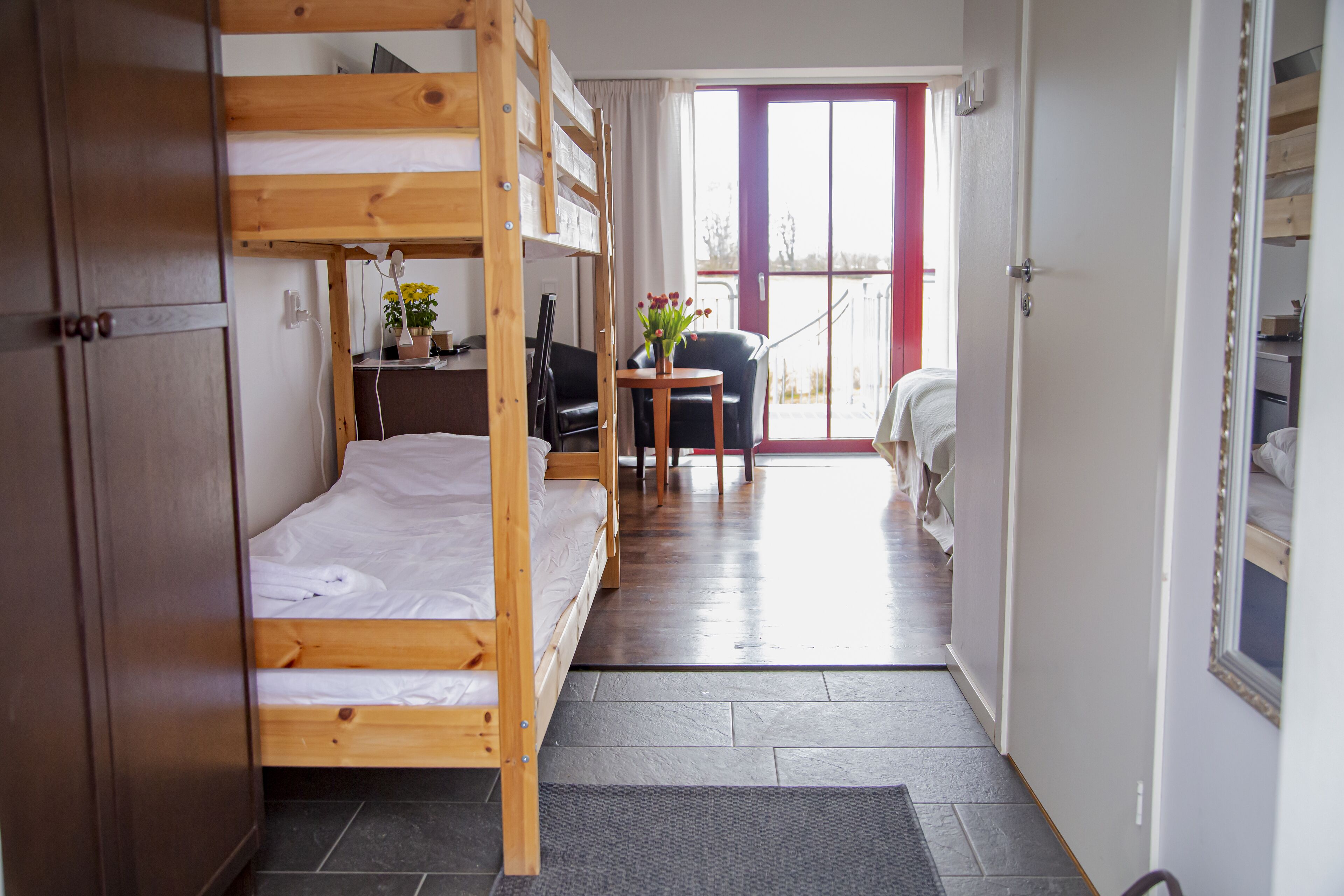 A room with bunk beds, a desk with a chair, a small table with flowers, and a view of the outside through a door.