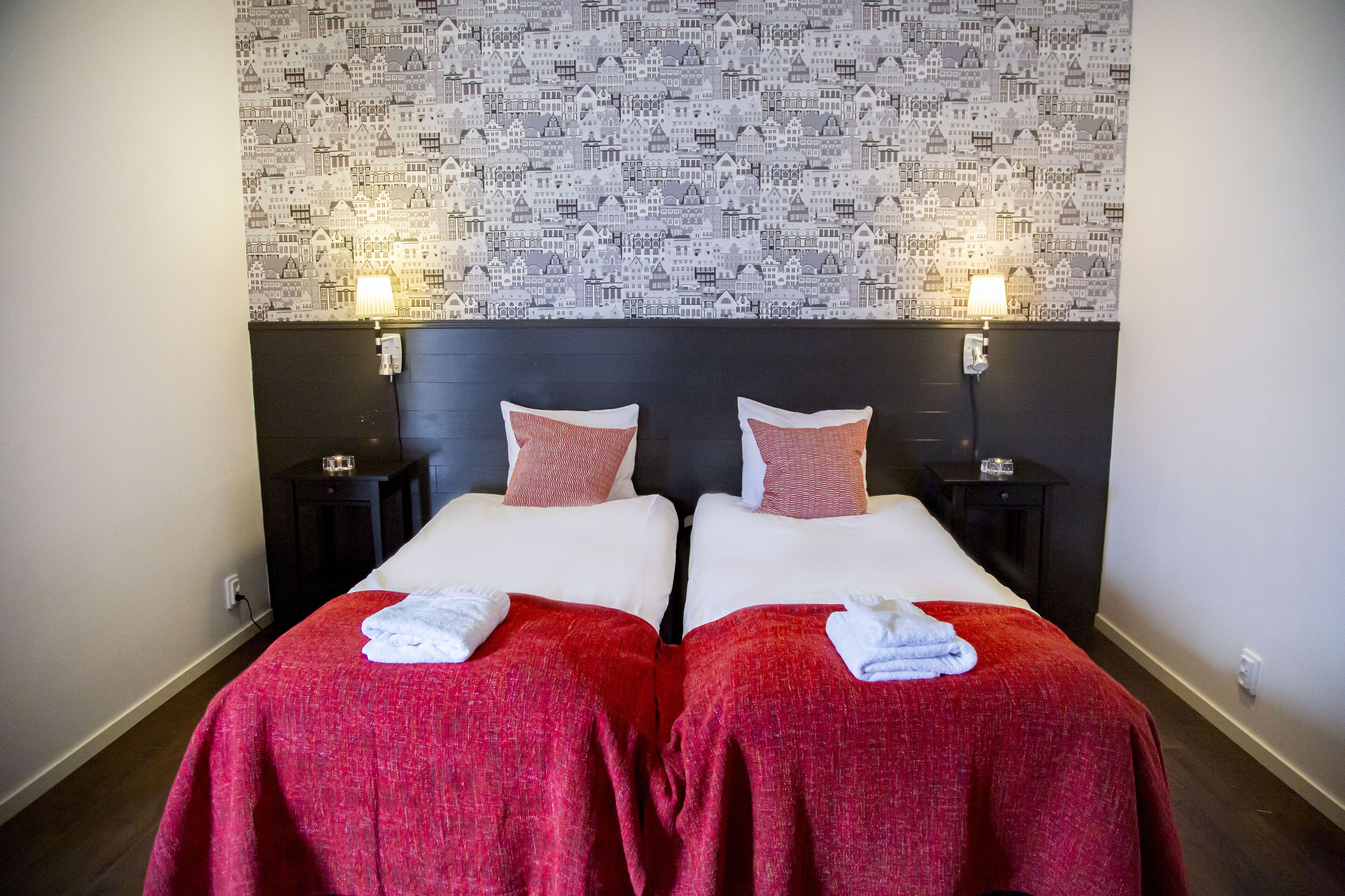 Ein Hotelzimmer mit zwei Betten, jeweils mit roter Bettdecke und weißen Kissen, und einem Wandpapier mit einem Muster von Gebäuden.