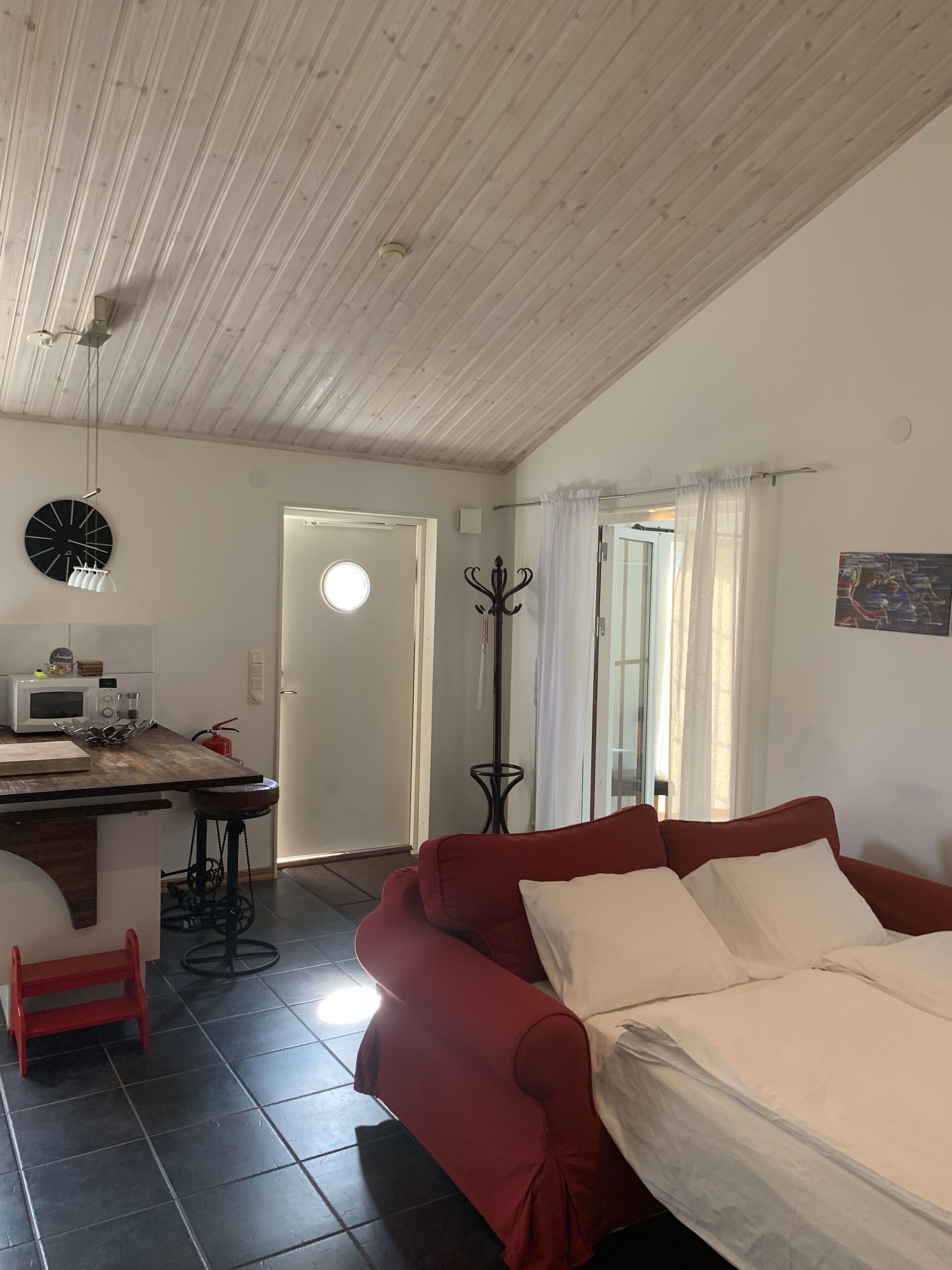 Una sala de estar acogedora con un sofá rojo, una mesa redonda, un microondas y un reloj en la pared.