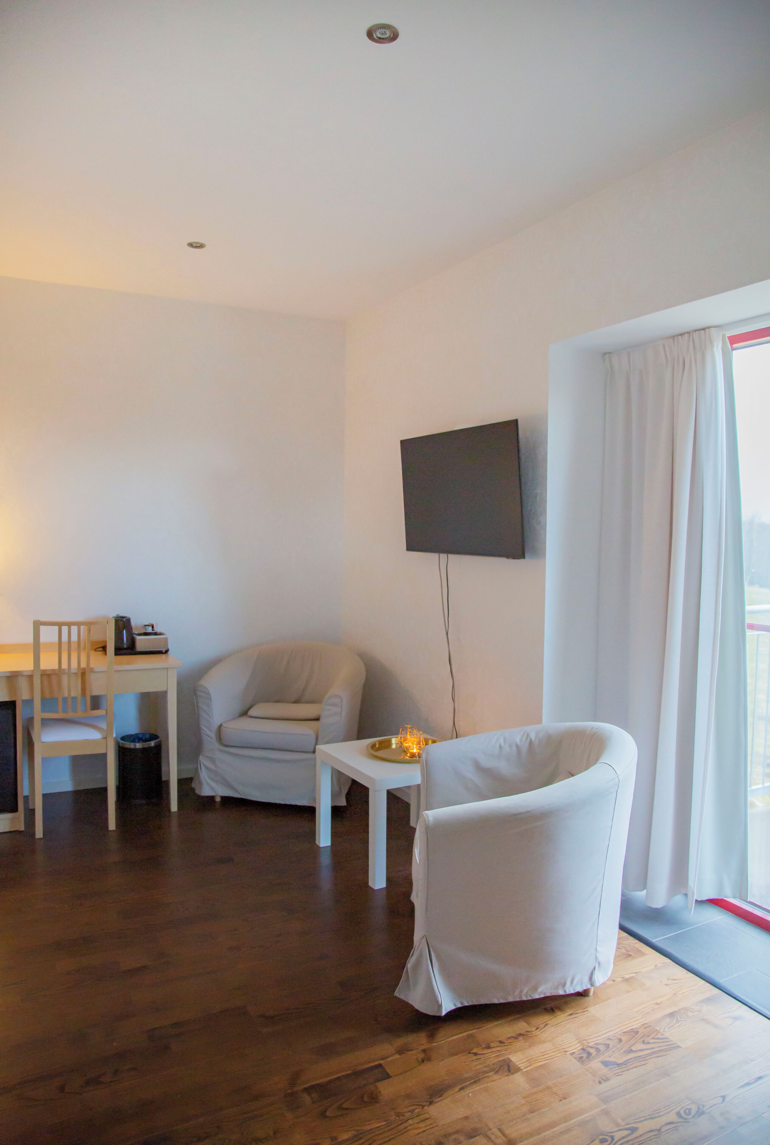 A room with a wooden floor, a desk with a chair, a television mounted on the wall, and a window with curtains.