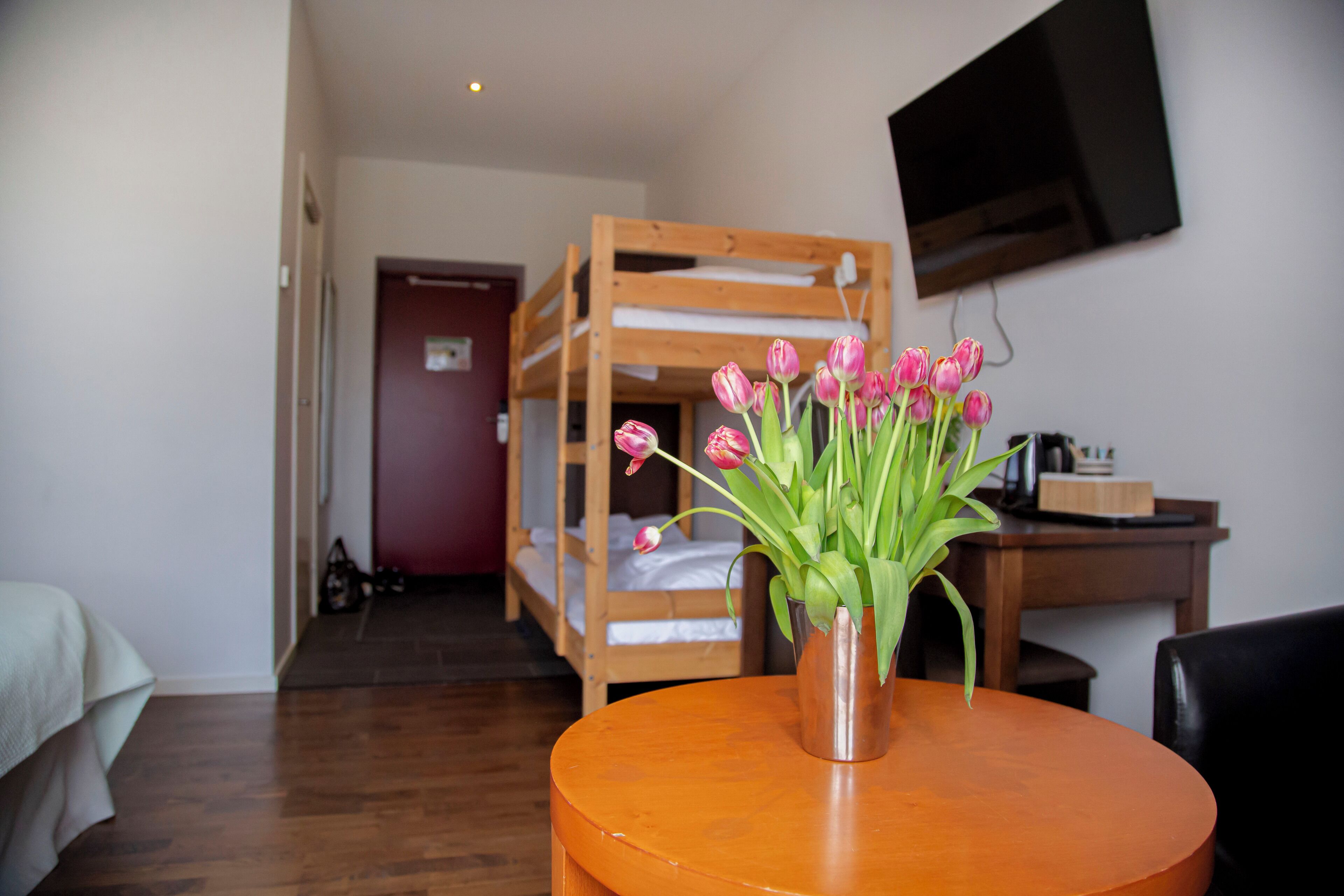 A room with a bunk bed, a round table with a vase of flowers, and a TV mounted on the wall.