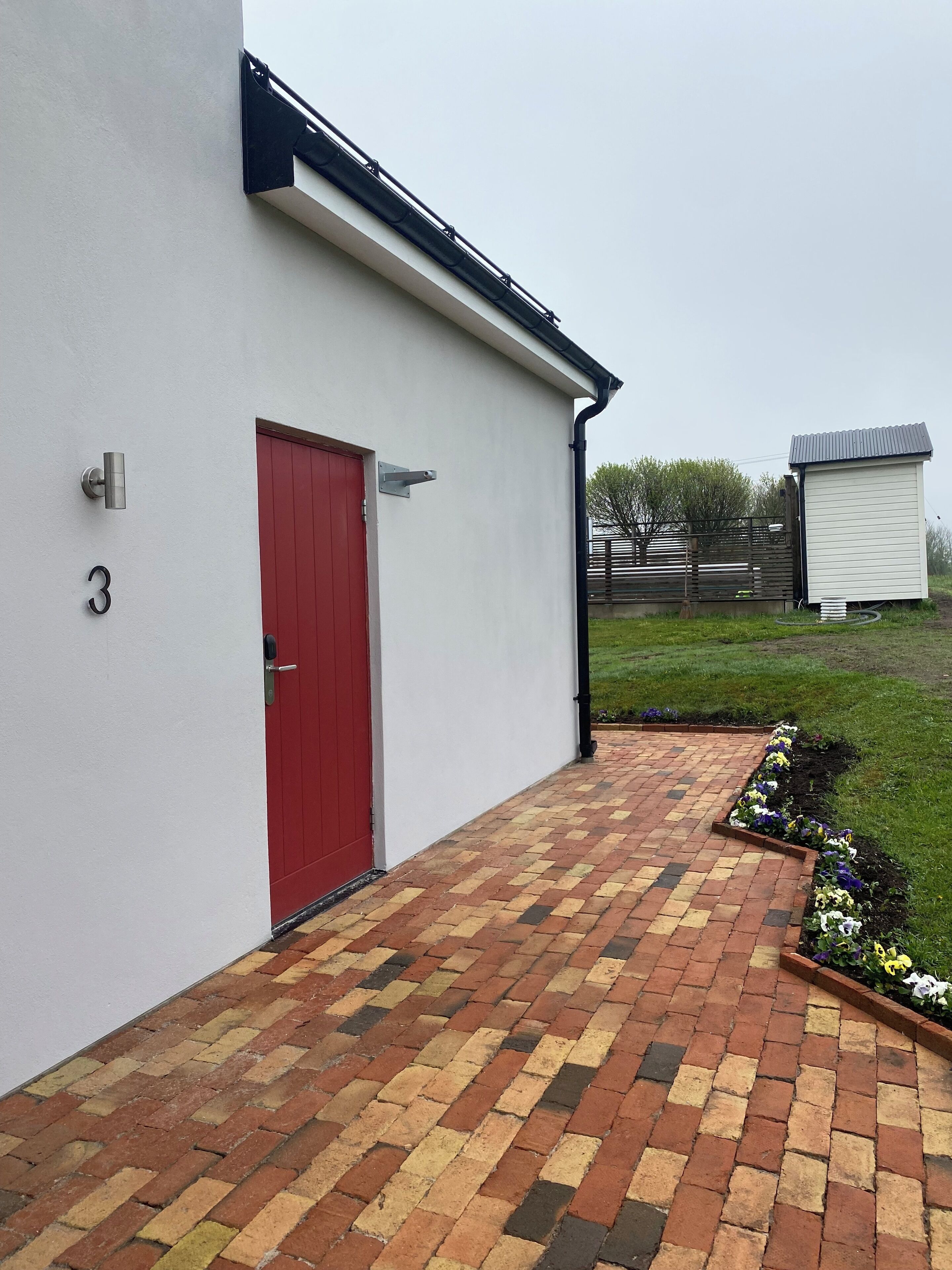 A building with a red door, a number '3' on the wall, and a paved walkway leading to a small shed.