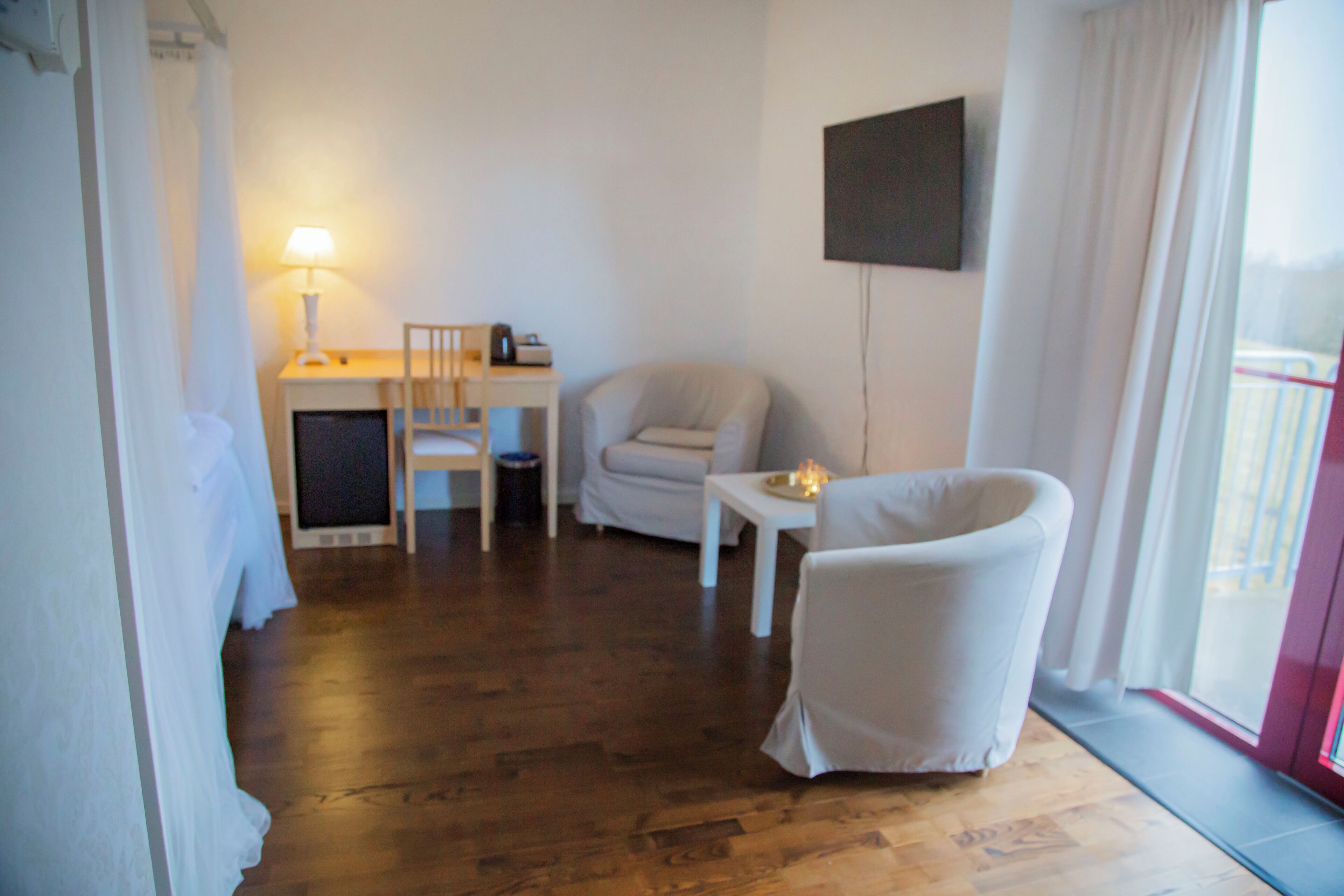 Una habitación con piso de madera, un sillón blanco, una mesita de noche con lámpara, un sofá y un televisor montado en la pared.