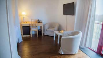 Une pièce avec un plancher en bois, un fauteuil blanc, une petite table avec une lampe, un canapé et un téléviseur fixé au mur.