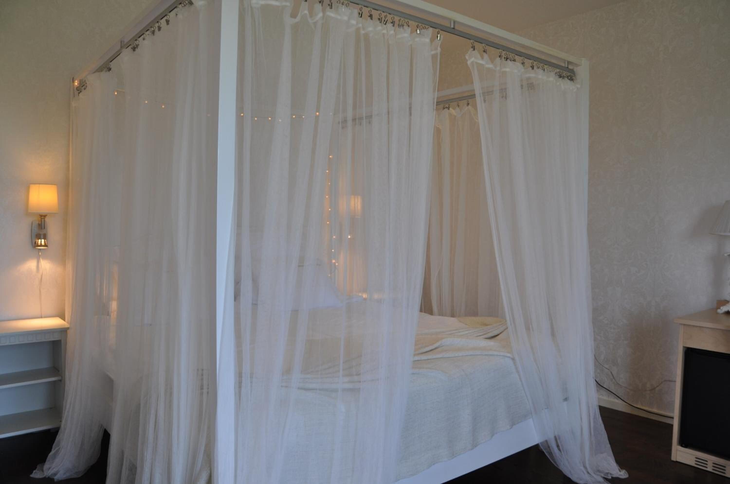 Una cama con dosel y cuatro columnas, con cubrecama blanco y una mesita de noche con lámpara.