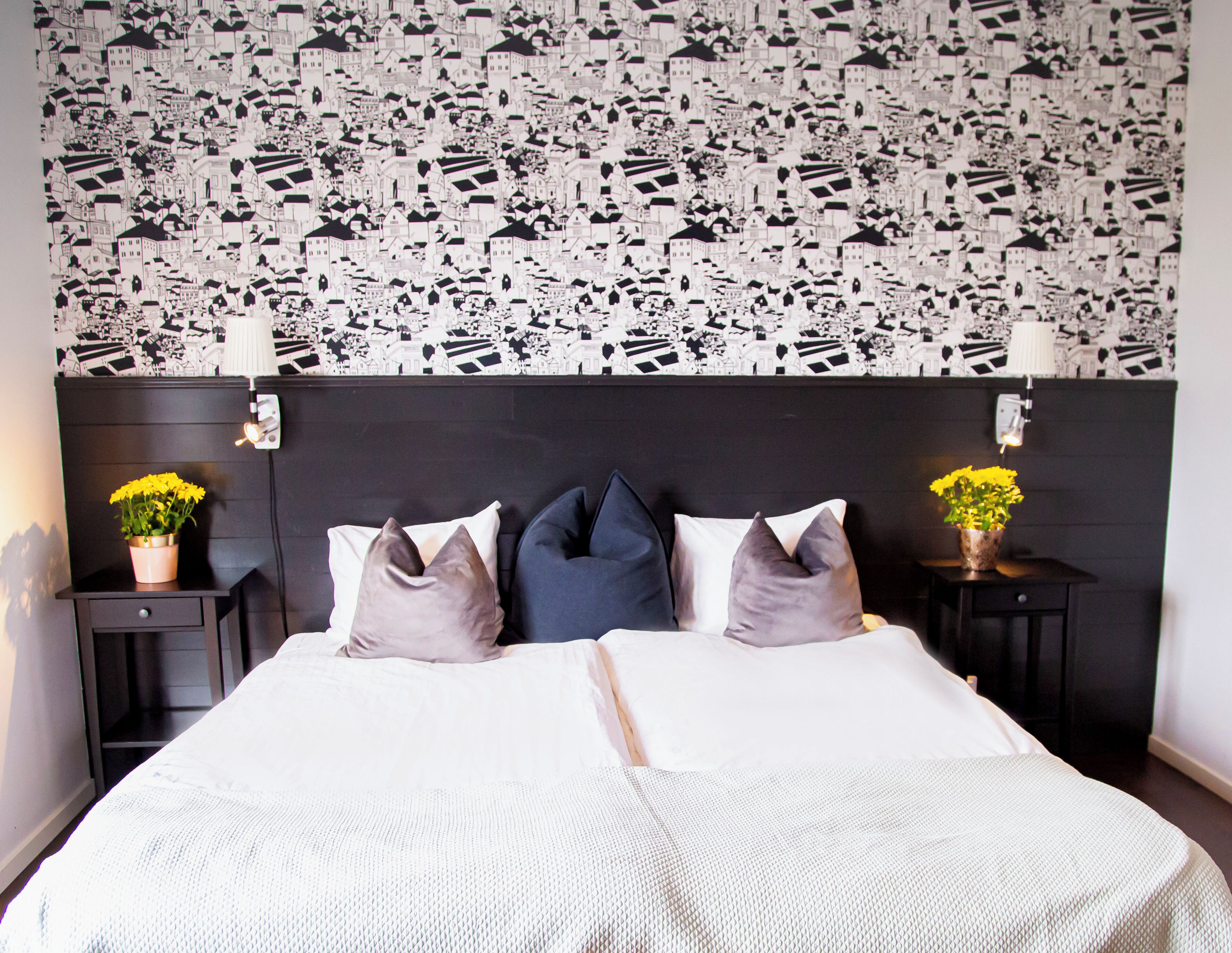 A bedroom with a patterned wallpaper, a bed with pillows, two bedside tables with lamps, and a nightstand with a flower vase.